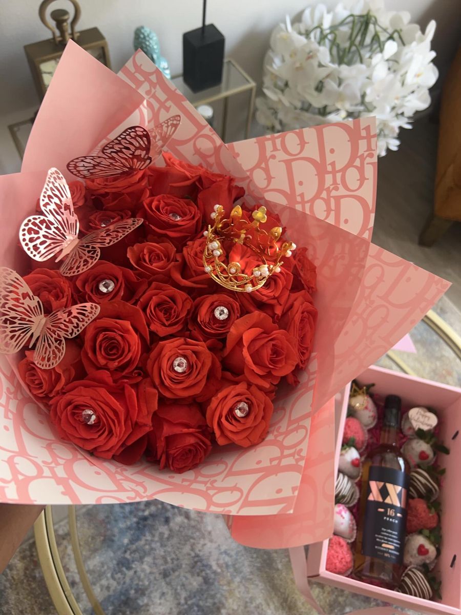 Red roses in pink wrap with crown and butterflies beside a box of strawberries and wine